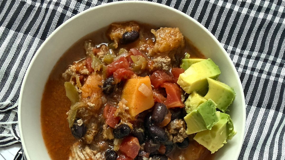 picture of bowl of soup with diced avocado on top in white bowl with black spoon and black and white fabric on white counter