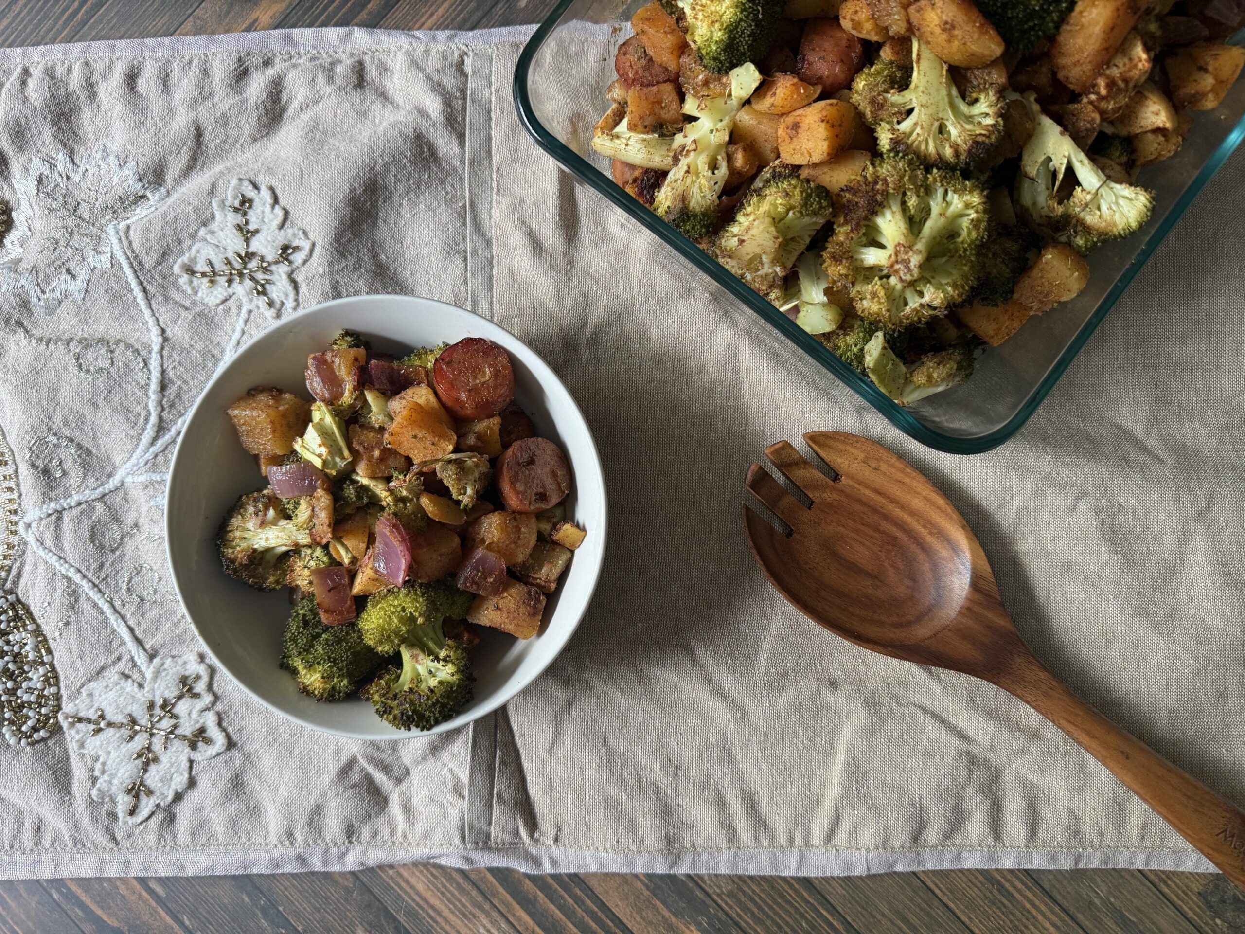 bowl of sheet pan meal of chicken sausage, butternut squash, broccoli, next to wooden spoon.